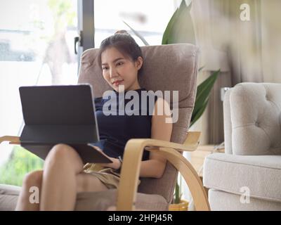Relaxed Asian young woman in comfortable outfits sits in her comfy armchair and works on her tablet. Stock Photo
