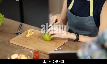 Close-up image, Woman with apron is preparing food in the kitchen, chopping green apple on wooden cutting board. Healthy lifestyle. Stock Photo