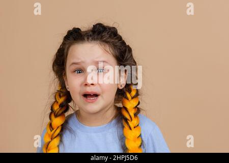 Sad small female with tears in eyes crying with open mouth with missing tooth with yellow kanekalon braids on head wearing light blue t-shirt on beige Stock Photo