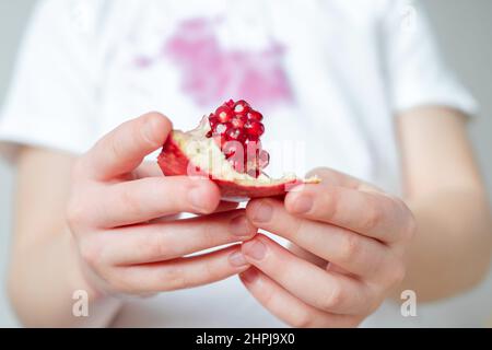 Close up slice of pomegranate seeds in the hands of a child. dirty stain of pomegranate juice after opening on clothes on background. High quality pho Stock Photo