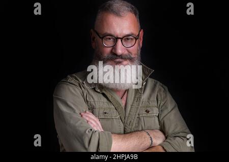 Mature bearded caucasian man looks slyly in camera with eyeglasses on his face, military style green shirt, black background Stock Photo