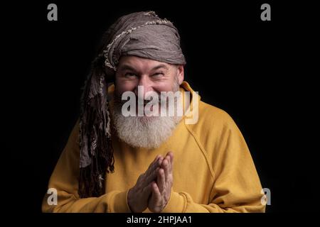 Mature bearded caucasian man looks slyly in camera and smile, gray scarf turban, black background Stock Photo