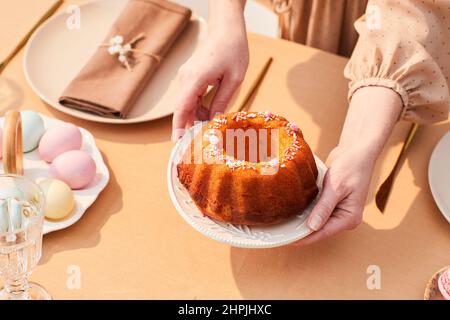 Close up of young woman putting cute homemade cake on dinner table decorated for Easter in sunlight, copy space Stock Photo