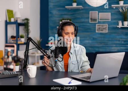 Social media influencer looking at laptop and recording video. Woman vlogger streaming online podcast on smartphone. Content creator in home studio using audio broadcasting equipment. Stock Photo