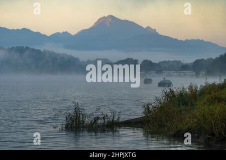 Shore of a lake in the early morning of autumn. The fog is clearing, allowing glimpses of moored boats. The lake water is choppy and a mountain can be seen with its peak illuminated by the first light Stock Photo