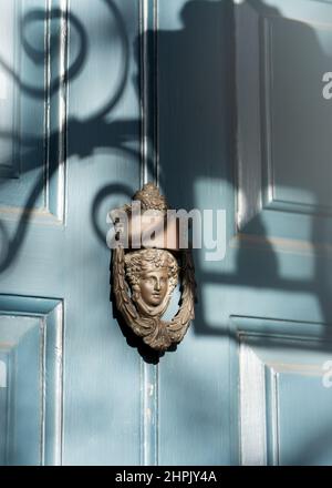 Ornate old brass door knocker with face on aqua blue wooden painted door. Shadow cast from street lamp on sunny day. Stock Photo