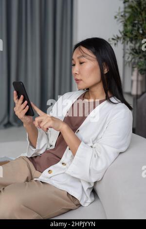 Young contemporary Asian female freelancer or employee scrolling in smartphone while sitting on sofa at home and working remotely Stock Photo
