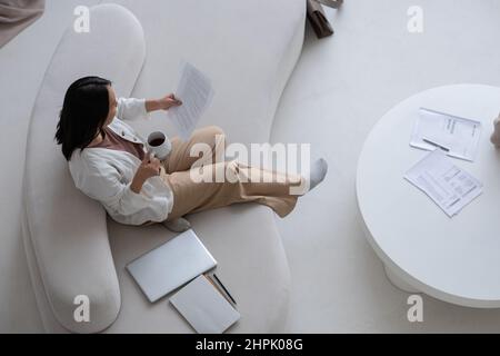 Young contemporary woman with drink sitting on large white comfortable sofa in living-room and looking through financial documents Stock Photo