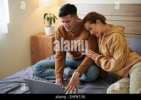 Young couple sitting together on the bed and using laptop to order things online Stock Photo