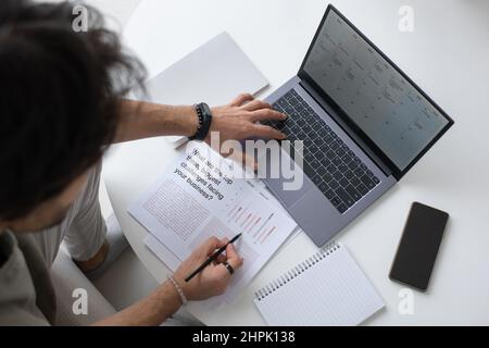 Contemporary auditor or analyst looking through text in document and analyzing financial data on laptop screen while working from home Stock Photo
