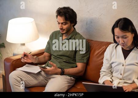 Two young intercultural employees in casualwear using wireless gadgets while sitting on leather couch and analyzing financial data Stock Photo