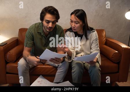 Two young confident intercultural employees with financial documents sitting on leather sofa and discussing and analyzing data Stock Photo