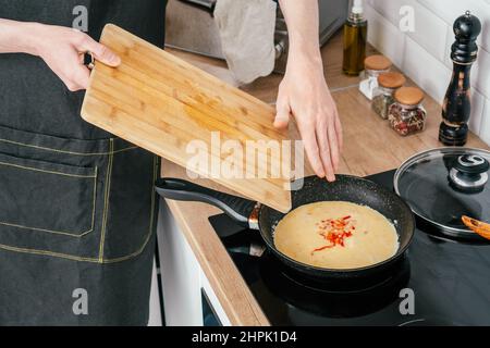 Unrecognizable man in black apron put chopped red bell pepper in omelette from wooden cutting board in frying pan on electric cooker near glass jars w Stock Photo