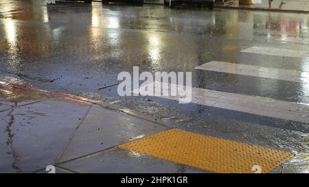 Rain drops on wet asphalt of city street in USA, raindrops falling on zebra crossroad. Cars lights reflection on road in rainy weather. Puddle of water by pavement. Night or twilight dusk atmosphere. Stock Photo