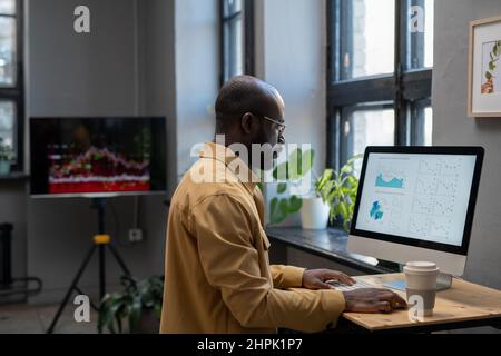 Young modern economist in casual clothes working with financial data in front of computer while sitting by desk in office Stock Photo