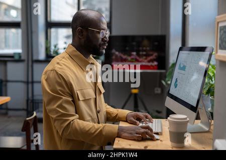 Contemporary young serious economist analyzing finnacial diagrams and graphs on computer screen while sitting by workplace in office Stock Photo