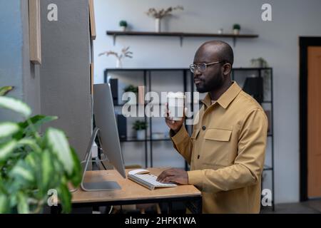 Young serious employee having drink and looking at data on computer screen while standing by workplace in urban office Stock Photo
