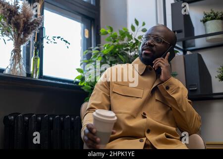 Contemporary young urban office worker with drink talking on smartphone while sitting in armchair against window and shelves Stock Photo
