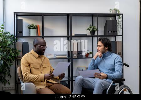 Small multi-ethnic group of businessmen with papers having discussion at working meeting while one of them sitting in wheelchair Stock Photo