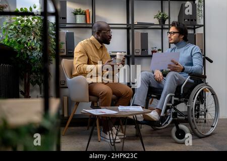 African-American businessman with drink communicating with his Caucasian colleague in wheelchair at working meeting in urban office Stock Photo