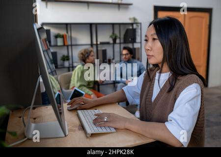 Young Asian businesswoman in casual clothes working in front of computer monitor on background of two intercultural coworkers Stock Photo