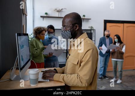 Young African-American economist analyzing data on computer screen while standing by desk in front of monitor with colleagues on background Stock Photo