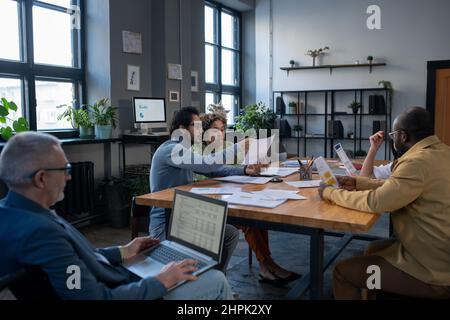 Group of intercultural economists discussing financial papers by large table while mature businessman with laptop preparing presentation Stock Photo