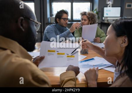 Asian businesswoman pointing at paper while discussing working points with colleague in front of two intercultural coworkers Stock Photo