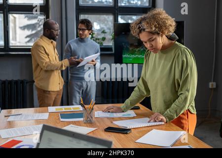Young female economist looking through financial data and analyzing it by table against two male colleagues preparing report Stock Photo