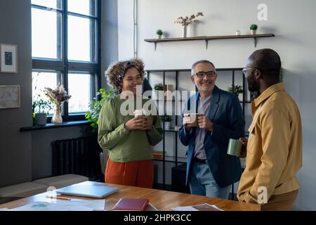 Three successful colleagues in smart casualwear having drinks at break by workplace in office while discussing new working points Stock Photo