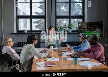 Contemporary young confident Asian business coach making presentation for group of intercultural coworkers at working meeting Stock Photo
