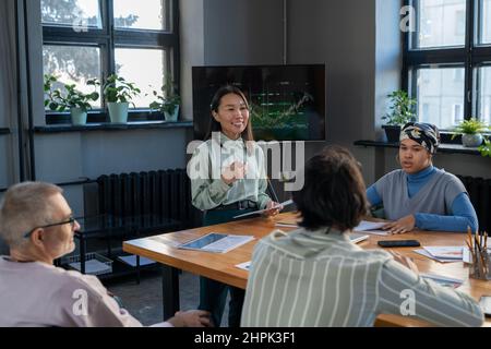 Young successful female Asian speaker communicating with audience at business training while standing by table with financial documents Stock Photo