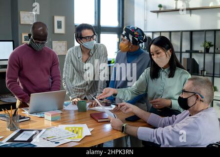 Contemporary intercultural employees in protective masks discussing financial data at meeting while gathered around table Stock Photo