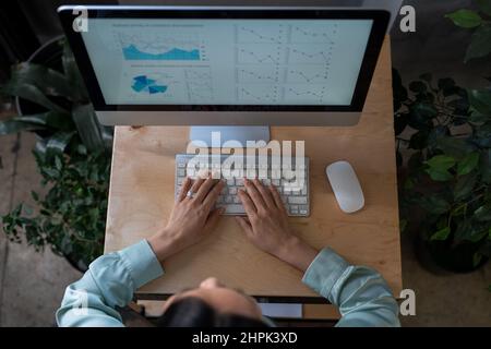 Overview of young female eonomist keeping her hands of computer keyboard while analyzing financial diagram and graphs Stock Photo