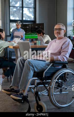 Mature employee with laptop on his knees sitting in wheelchair in front of camera against group of young intercultural colleagues Stock Photo