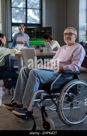 Mature employee with laptop on his knees sitting in wheelchair in front of camera against group of young intercultural colleagues Stock Photo