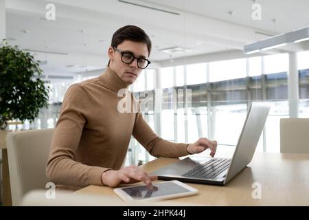 Young serious high-tech developer using tablet and laptop while sitting by tabel and working over new programming project Stock Photo