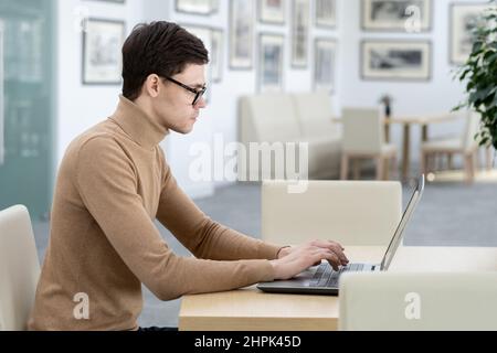 Side view of young serious developer of technology company typing on laptop keyboard while sitting by workplace in modern office Stock Photo