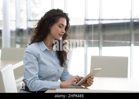 Pretty brunette female manager in formalwear touching screen of tablet while analyzing online data by workplace in large modern office Stock Photo