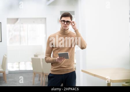 Young contemporary ceo of large high tech corporation with digital tablet keeping fingers on eyeglasses while looking at camera Stock Photo