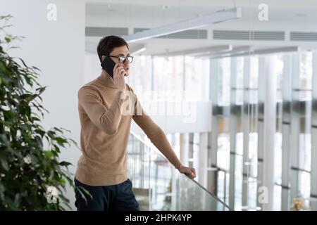 Young confident ceo in smart casualwear standing in office building and holding mobile phone by ear while consulting client of company Stock Photo