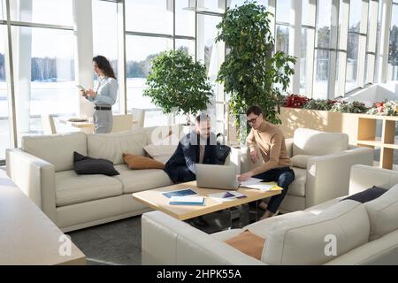 Two confident businessmen sitting by table in front of laptop while discussing online data and new business strategies at meeting Stock Photo