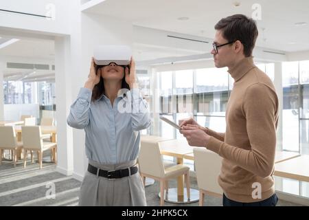 Young businessman with tablet standing by female colleague in vr goggle and entering data in digital database during work over project Stock Photo