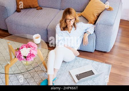 Full length shot of middle aged woman sitting in the living room and using her laptop while having online meeting. Home office. Stock Photo
