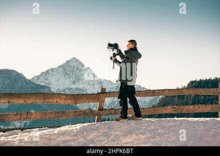 Videographer man recording video using gimbal stabilizer equipment and camera. Snow and mountains in winter background.