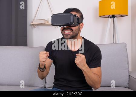 Young man in VR glasses headset at home on sofa excited gesturing, future technology, education video gaming Stock Photo