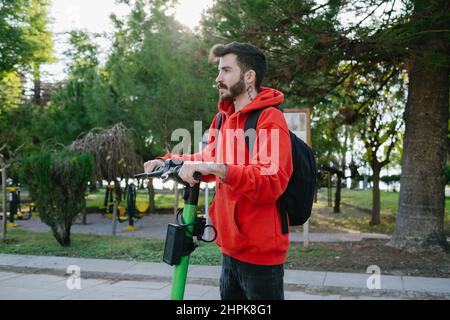 Driving electric scooter, young attractive student with backpack is driving his scooter at bright sunny day park background. Stock Photo