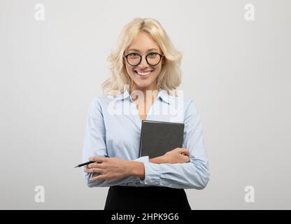 Portrait of happy female entrepreneur in formal wear holding notepad and pen, posing over ligth grey studio background Stock Photo