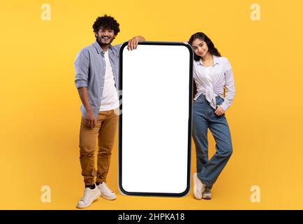 Excited young indian man and woman leaning on giant smartphone with mockup, promoting app or website Stock Photo