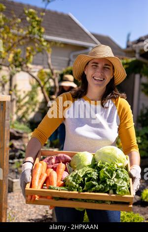 Portrait of smiling caucasian mature woman with vegetables in wooden crate standing in backyard Stock Photo